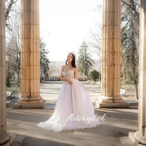 May include: A woman wearing a white strapless wedding dress with a beaded bodice and a tulle skirt. She is standing in a white stone archway with trees in the background. The text "MatchingLook" is visible in the bottom right corner of the image.