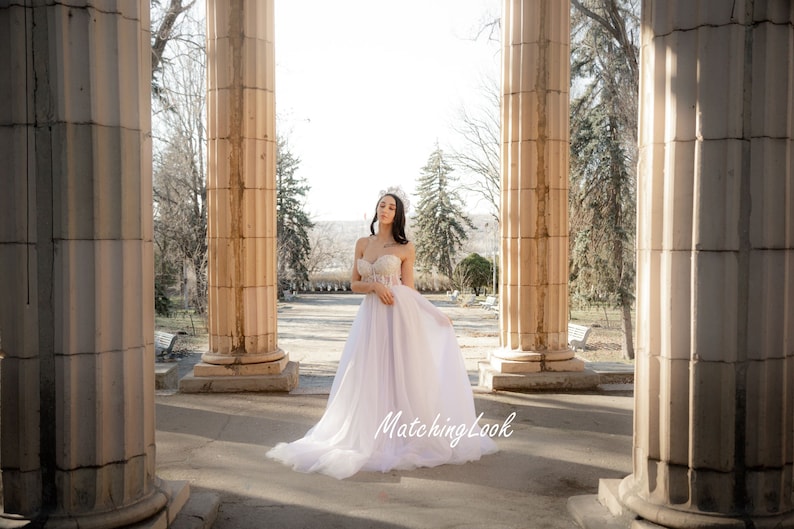 May include: A woman wearing a white strapless wedding dress with a beaded bodice and a tulle skirt. She is standing in a white stone archway with trees in the background. The text "MatchingLook" is visible in the bottom right corner of the image.