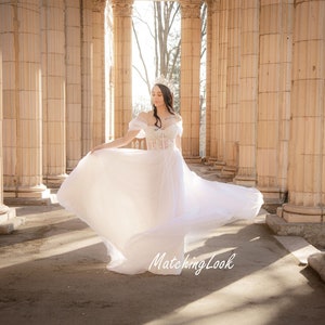 May include: A woman in a white wedding dress with a flowing skirt stands in a colonnade. She is wearing a tiara and has her hair pulled back. The dress has a lace bodice and a tulle skirt. The woman is smiling and looking down at the ground.