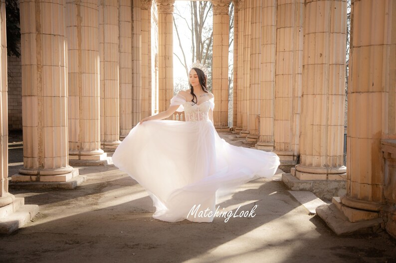May include: A woman in a white wedding dress with a flowing skirt stands in a colonnade. She is wearing a tiara and has her hair pulled back. The dress has a lace bodice and a tulle skirt. The woman is smiling and looking down at the ground.