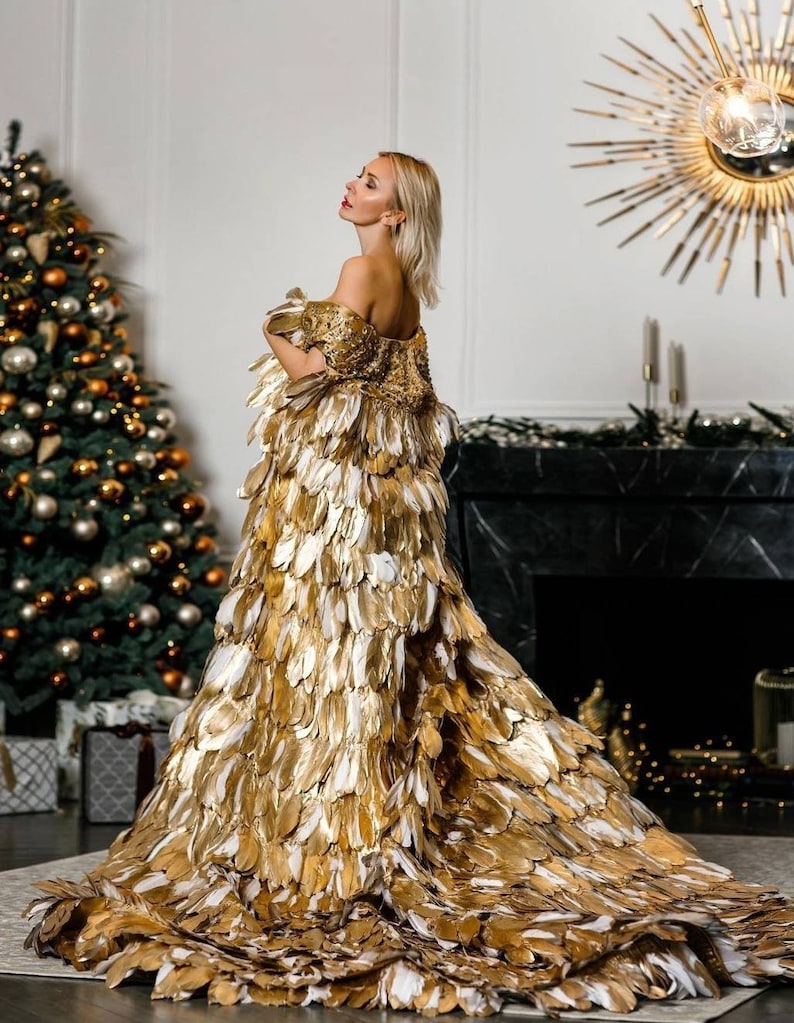 May include: A woman wearing a gold and white feather gown. The gown has a long train and is made of many feathers. The woman is standing in a room with a Christmas tree and a fireplace.