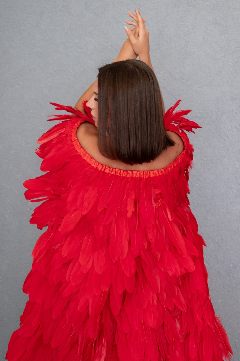 May include: A red feather cape with a ruffled neckline. The cape is made of many layers of feathers and is worn by a person with long brown hair.