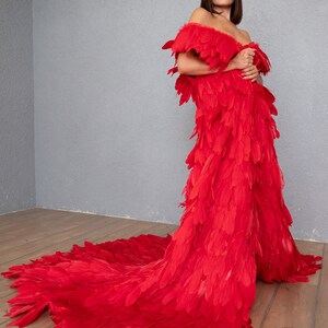 May include: A woman wearing a long, red dress made entirely of feathers. The dress has an off-the-shoulder neckline and a flowing train.