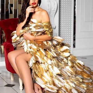 May include: A woman wearing a gold and white feather dress with a red lip and red high heels. She is sitting on a red chair in a white room.