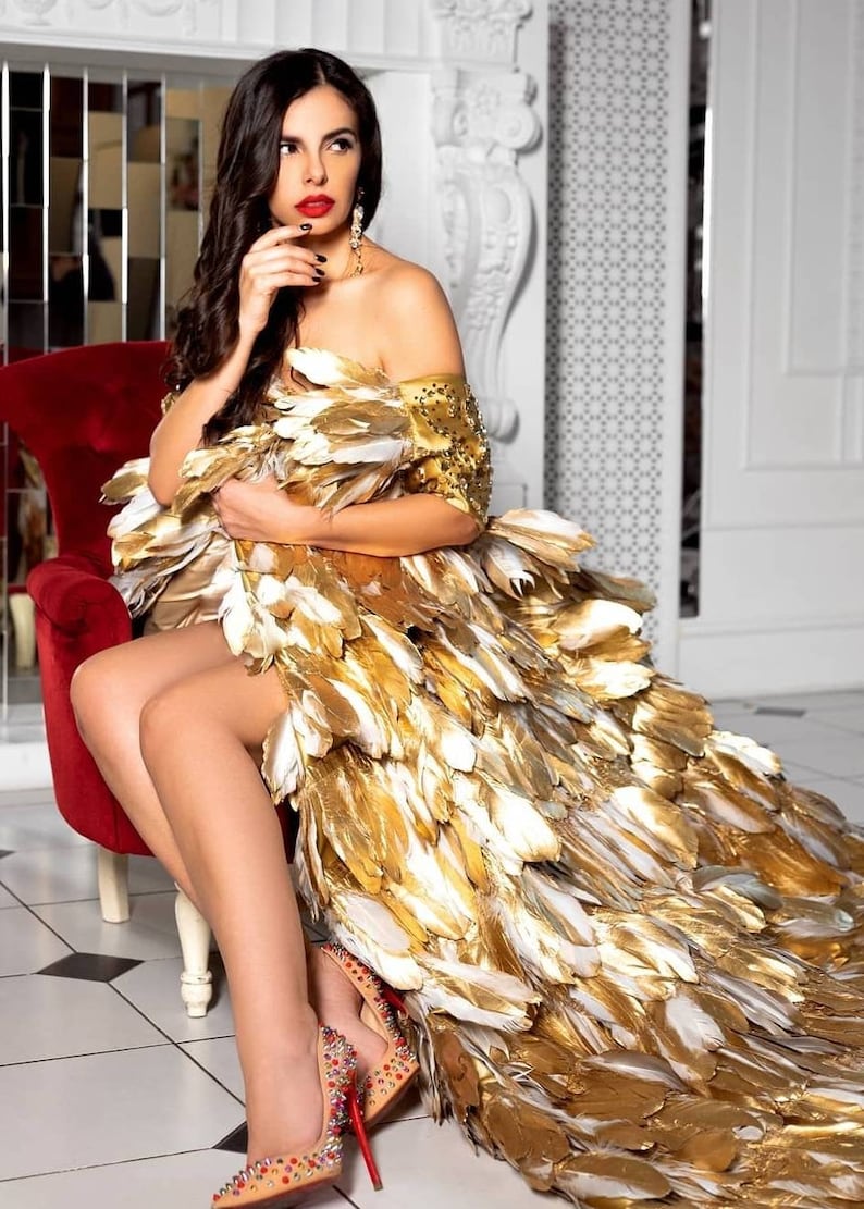 May include: A woman wearing a gold and white feather dress with a red lip and red high heels. She is sitting on a red chair in a white room.