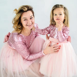 May include: A mother and daughter wearing matching pink sequin dresses with tulle skirts. The dresses have a bow on the back. The mother is smiling and looking at the camera. The daughter is looking at the camera with a serious expression.