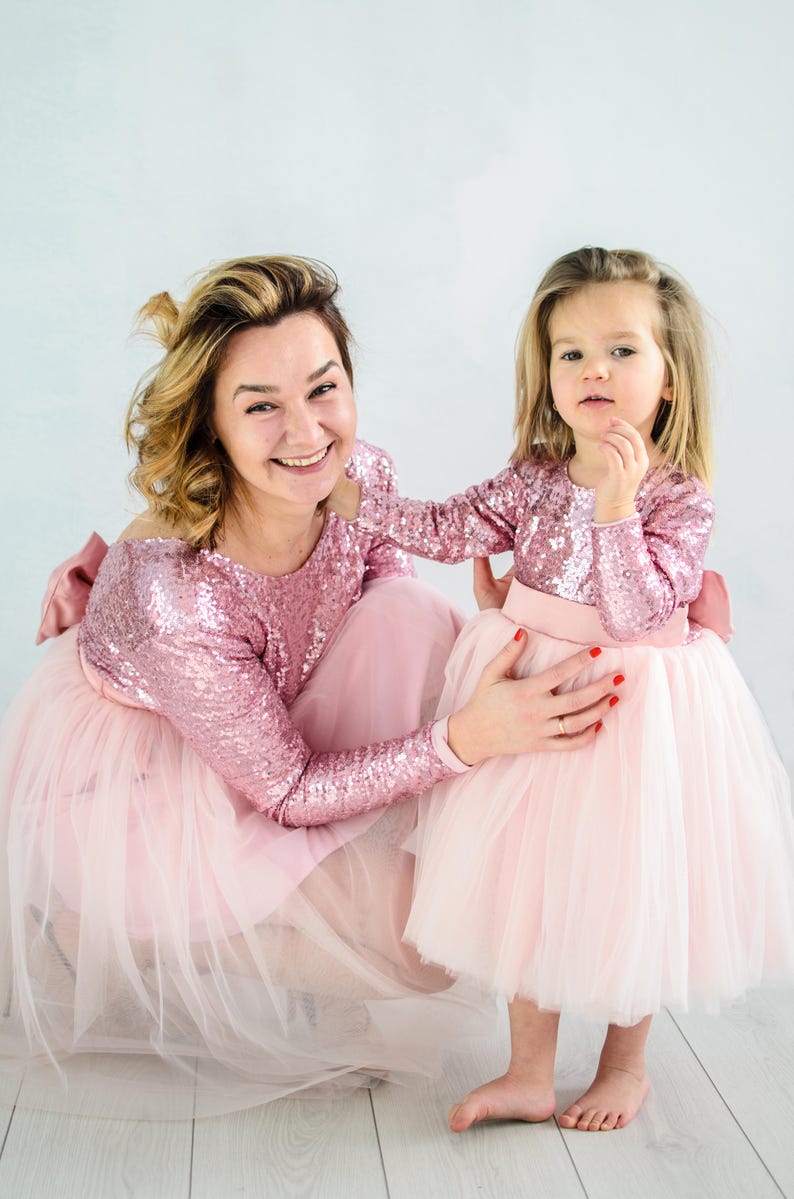 May include: A mother and daughter wearing matching pink sequin dresses with tulle skirts. The dresses have a bow on the back. The mother is smiling and looking at the camera. The daughter is looking at the camera with a serious expression.