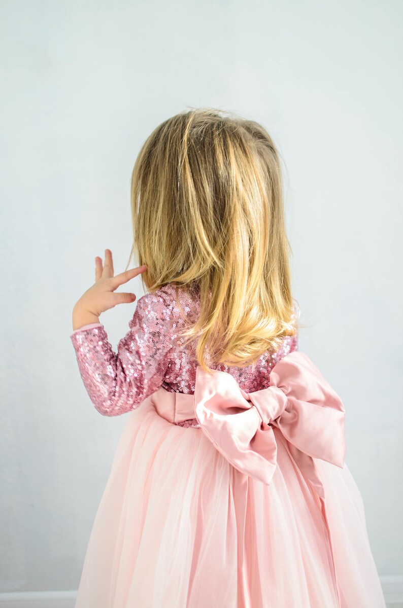 May include: A young girl wearing a pink tulle dress with a large pink satin bow on the back. The dress has a sequined bodice.