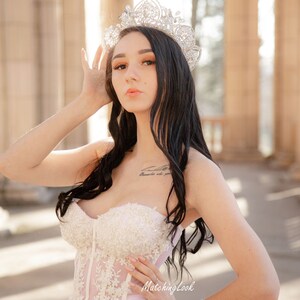 May include: A woman wearing a white strapless gown with lace detailing and a sparkling tiara. The gown has a tulle skirt and a fitted bodice. The woman is standing in front of a stone archway.