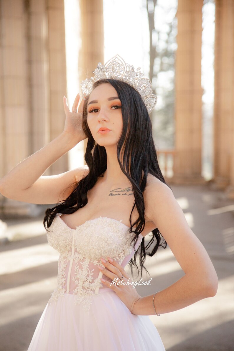 May include: A woman wearing a white strapless gown with lace detailing and a sparkling tiara. The gown has a tulle skirt and a fitted bodice. The woman is standing in front of a stone archway.