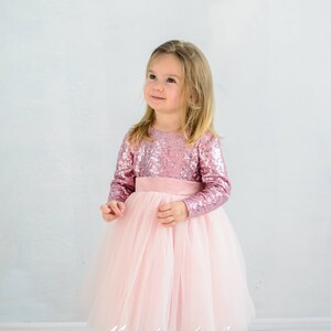 May include: A young girl wearing a pink long-sleeved sequined dress with a pink tulle skirt and a matching pink satin sash. The dress has a round neckline and long sleeves. The girl is standing on a light wood floor.