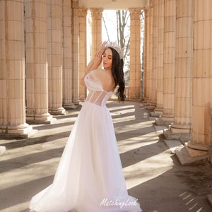May include: A woman wearing a white tulle wedding dress with a beaded bodice and a crown stands in a colonnade. The dress has a sweetheart neckline and off-the-shoulder straps. The woman is looking down at her hands.