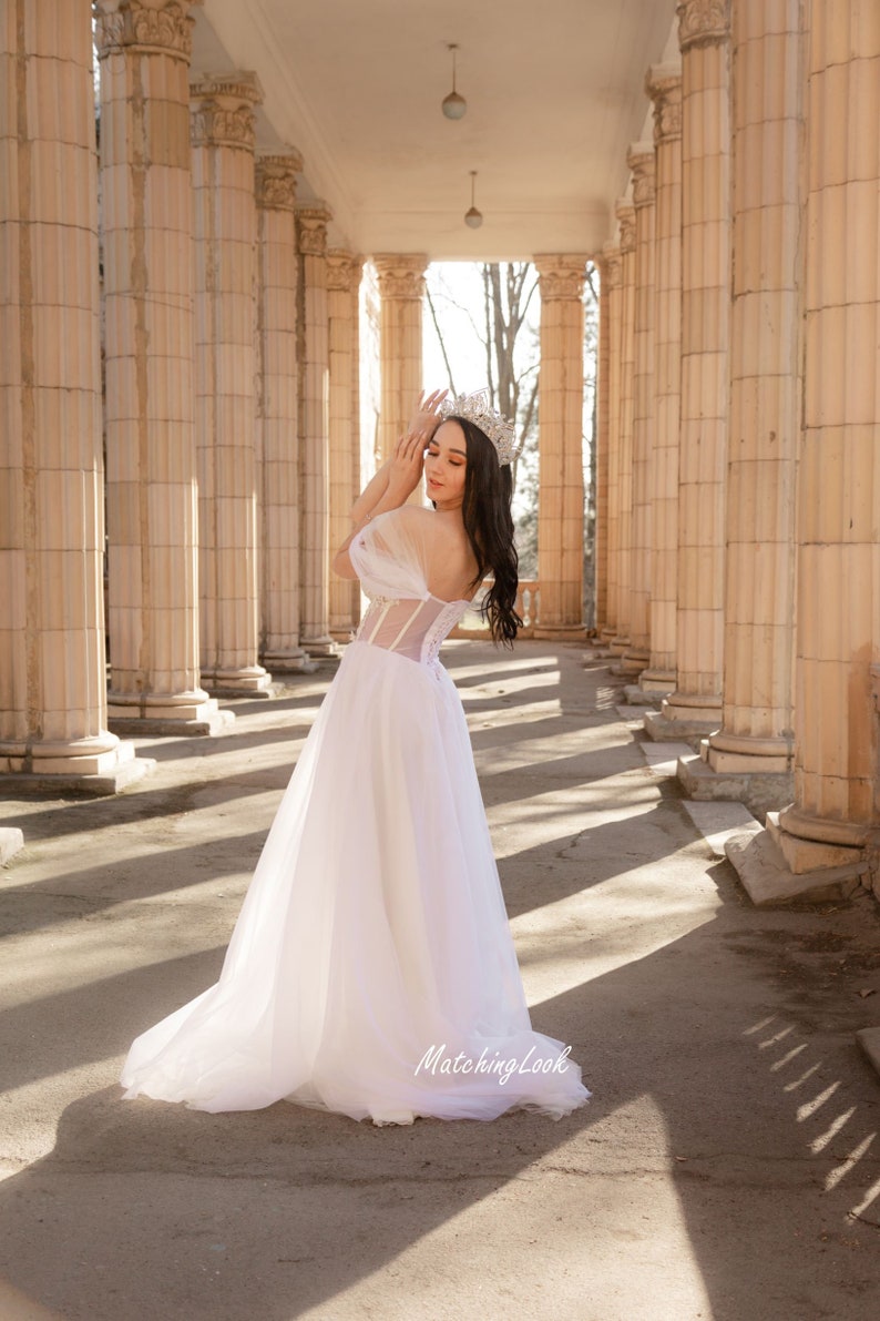 May include: A woman wearing a white tulle wedding dress with a beaded bodice and a crown stands in a colonnade. The dress has a sweetheart neckline and off-the-shoulder straps. The woman is looking down at her hands.