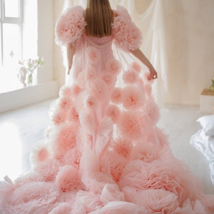 May include: A light pink, floor-length gown with a voluminous design. The gown features large, ruffled floral accents on the shoulders and cascading down the skirt. The fabric appears soft and flowing, creating a dreamy aesthetic.