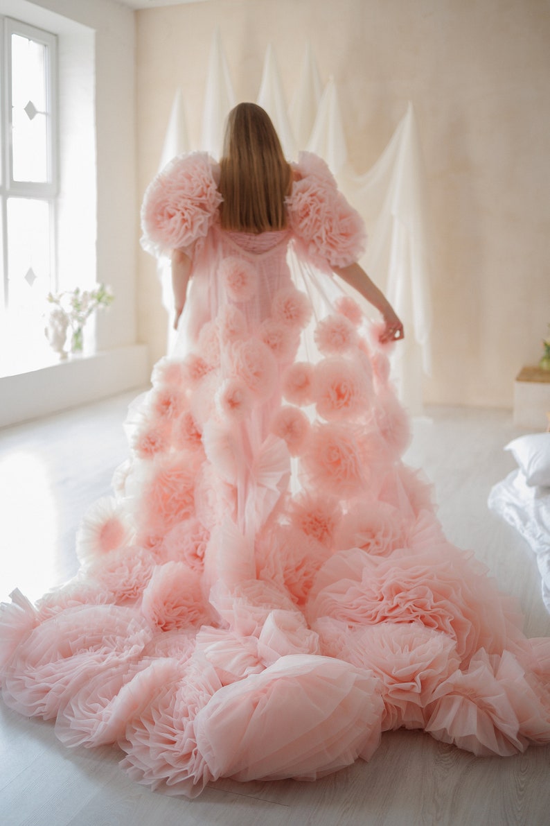 May include: A light pink, floor-length gown with a voluminous design. The gown features large, ruffled floral accents on the shoulders and cascading down the skirt. The fabric appears soft and flowing, creating a dreamy aesthetic.