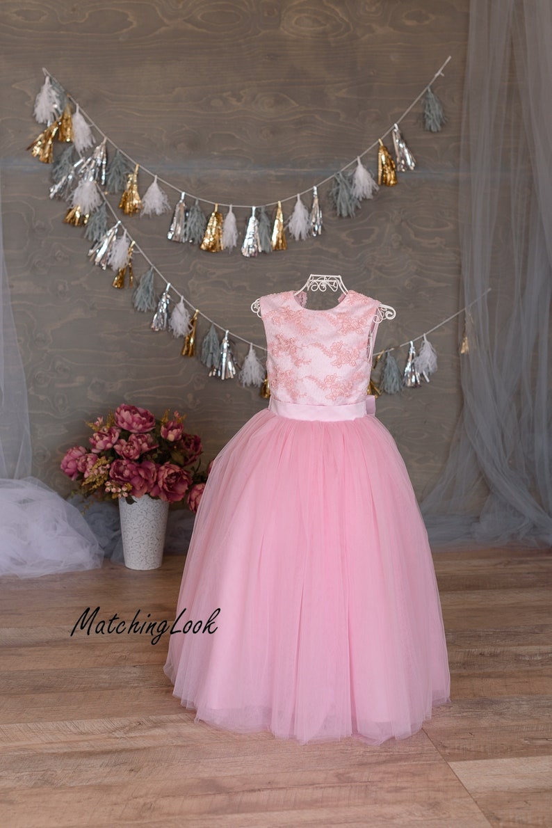 Puede incluir: Un vestido de tul rosa con un corpi&ntilde;o de encaje y una faja de sat&eacute;n. El vestido es sin mangas y tiene una falda completa. El vestido est&aacute; en un maniqu&iacute; blanco y est&aacute; frente a un fondo de madera.