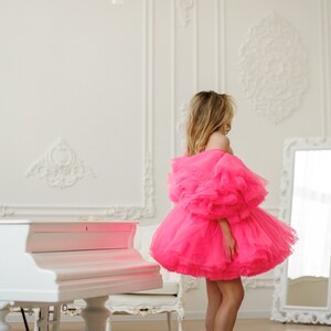 May include: A woman wearing a bright pink tulle dress with ruffled sleeves and a white grand piano in the background.