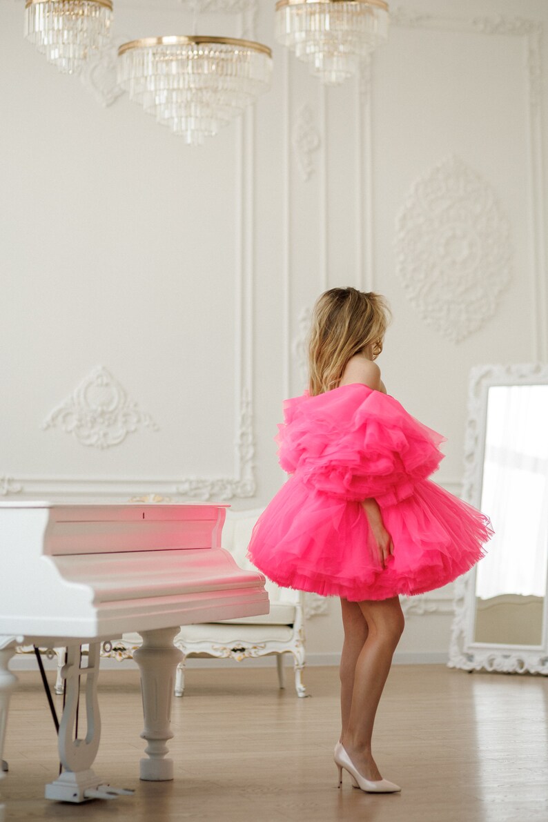 May include: A woman wearing a bright pink tulle dress with ruffled sleeves and a white grand piano in the background.