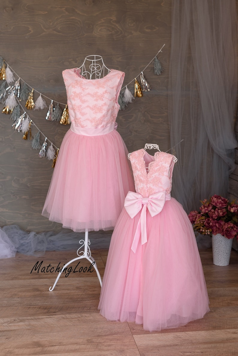 Puede incluir: Dos vestidos de tul rosa con corpi&ntilde;os de encaje. El vestido m&aacute;s grande tiene un lazo de sat&eacute;n en la espalda. Los vestidos est&aacute;n en un soporte blanco sobre un fondo de madera.