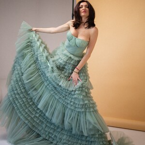 Pu&ograve; includere: Una donna indossa un abito di tulle verde salvia con pi&ugrave; strati di balze. L'abito &egrave; senza spalline e ha un corpetto aderente. La donna &egrave; in piedi davanti a uno sfondo marrone chiaro.