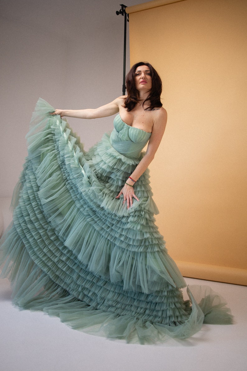 Pu&ograve; includere: Una donna indossa un abito di tulle verde salvia con pi&ugrave; strati di balze. L'abito &egrave; senza spalline e ha un corpetto aderente. La donna &egrave; in piedi davanti a uno sfondo marrone chiaro.