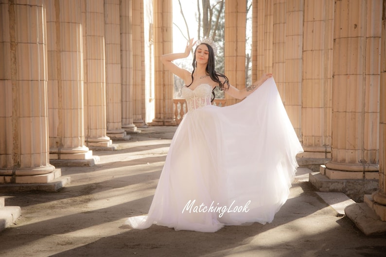 May include: A woman wearing a white wedding dress with a lace bodice and a flowing skirt. She is standing in a colonnade with sunlight streaming in.