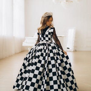 Könnte beinhalten: Ein junger Mensch trägt ein schwarz-weiß kariertes Ballkleid mit einem silbernen Diadem. Das Kleid hat ein eng anliegendes Oberteil und einen weiten Rock. Die Ärmel sind aus schwarzer Spitze. Der Hintergrund ist ein weißer Raum mit einem Klavier.