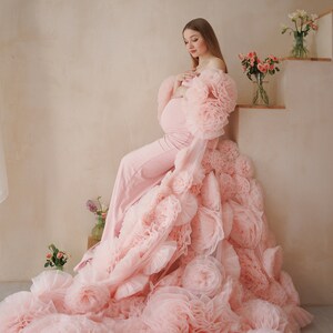 May include: A light pink maternity gown with a dramatic, layered floral design. The gown features a fitted jumpsuit and a flowing train of large, ruffled fabric flowers. The model is seated near a staircase with floral arrangements.