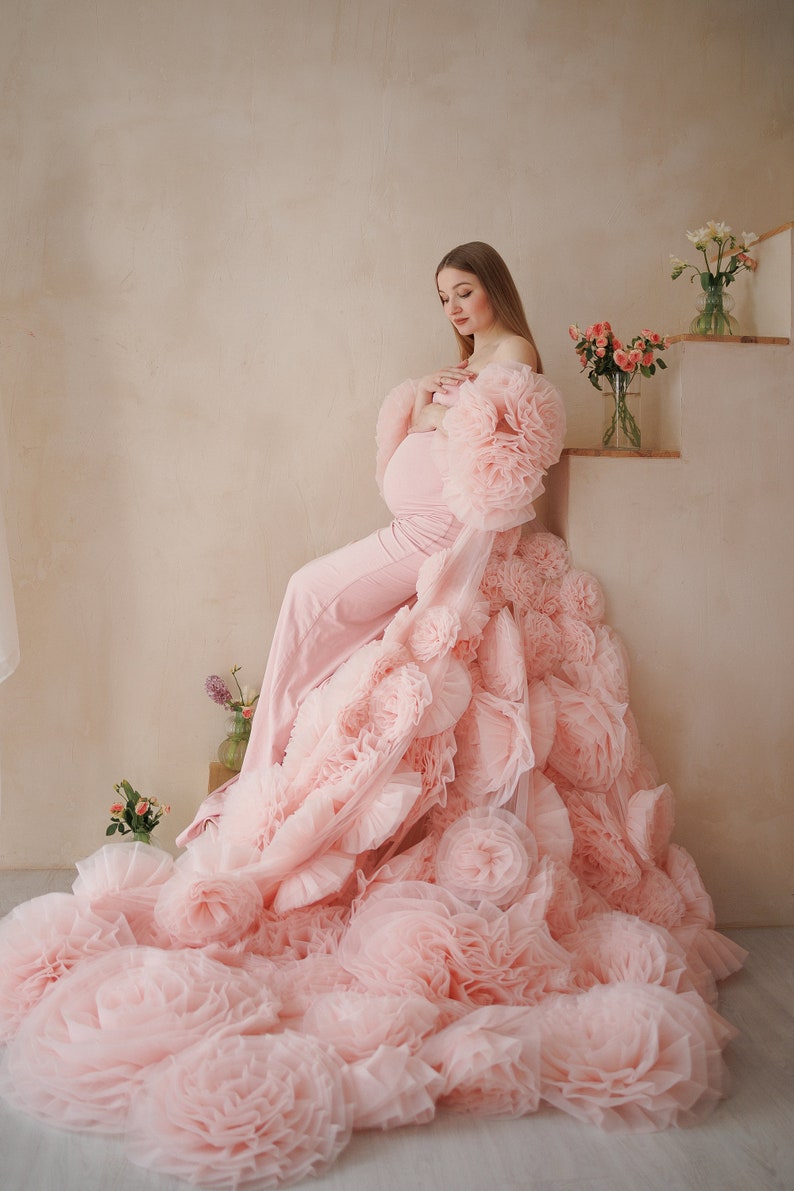 May include: A light pink maternity gown with a dramatic, layered floral design. The gown features a fitted jumpsuit and a flowing train of large, ruffled fabric flowers. The model is seated near a staircase with floral arrangements.