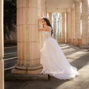 May include: A woman in a white wedding dress with a tulle skirt stands in front of a column. She is wearing a tiara and has her hair pulled back. The background is a colonnade with trees visible through the columns.