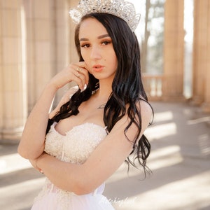 May include: A woman wearing a white strapless gown with a beaded bodice and a sparkling tiara. She is standing in front of a stone archway with columns.