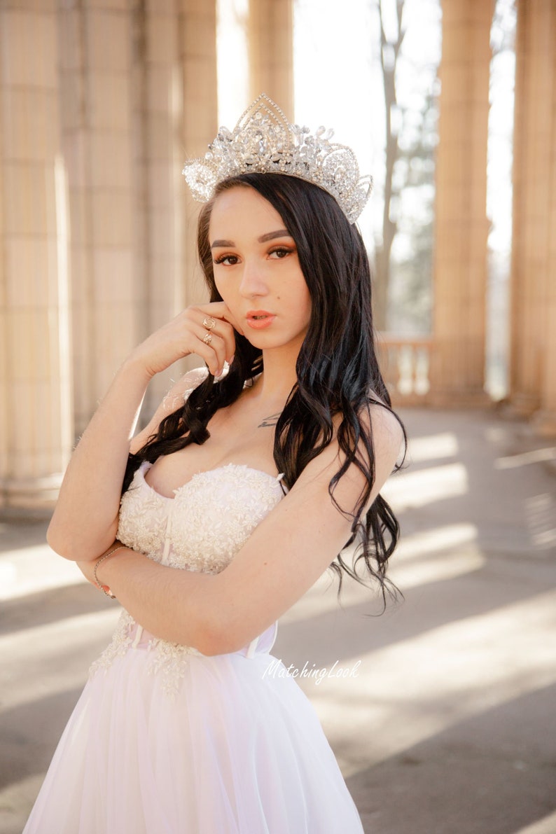 May include: A woman wearing a white strapless gown with a beaded bodice and a sparkling tiara. She is standing in front of a stone archway with columns.