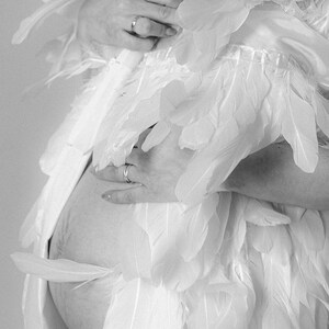 May include: A pregnant person wearing a white dress with feathers. The person is holding their belly with one hand and has a ring on their finger.