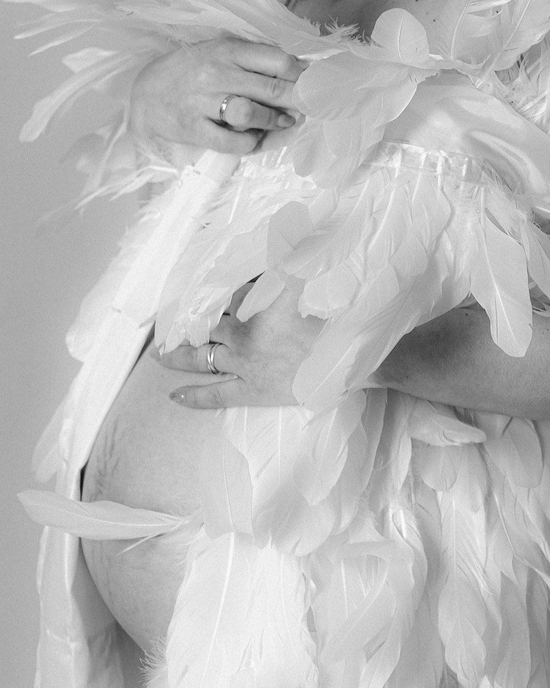 May include: A pregnant person wearing a white dress with feathers. The person is holding their belly with one hand and has a ring on their finger.