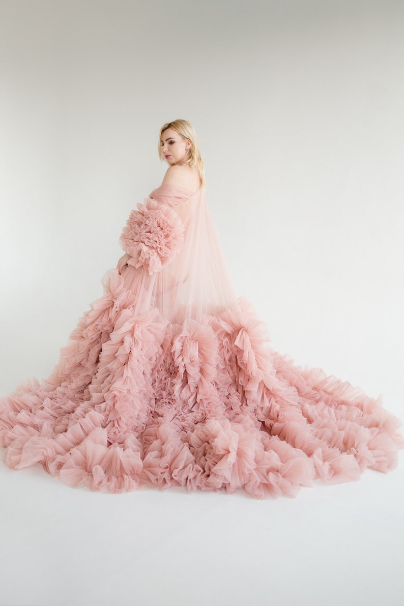 Pu&ograve; includere: Una donna indossa un lungo abito di tulle rosa con balze. L'abito ha una gonna fluida e un corpetto aderente. La donna &egrave; in piedi su uno sfondo bianco.