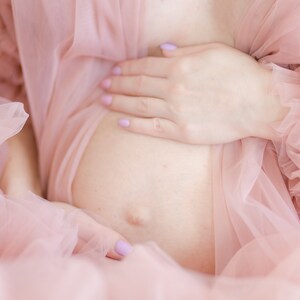 Pu&ograve; includere: Una persona incinta che indossa un abito di tulle rosa con volant. La persona tiene la pancia con una mano.