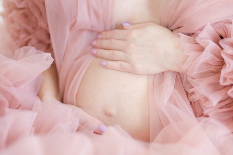 Pu&ograve; includere: Una persona incinta che indossa un abito di tulle rosa con volant. La persona tiene la pancia con una mano.