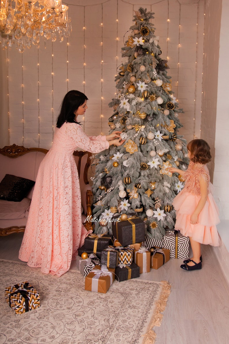 Puede incluir: Una mujer y una ni&ntilde;a peque&ntilde;a decoran un &aacute;rbol de Navidad con adornos blancos y dorados. El &aacute;rbol est&aacute; decorado con poinsettias blancas y adornos rayados dorados y negros. Hay regalos envueltos en papel de regalo negro, blanco y dorado en la base del &aacute;rbol.