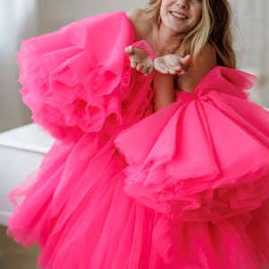 May include: A woman wearing a bright pink tulle dress with multiple layers and ruffles. She is smiling and blowing a kiss.