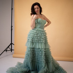 Pu&ograve; includere: Una donna indossa un abito di tulle verde salvia con pi&ugrave; strati di balze. L'abito ha un corpetto senza spalline e una gonna fluida.