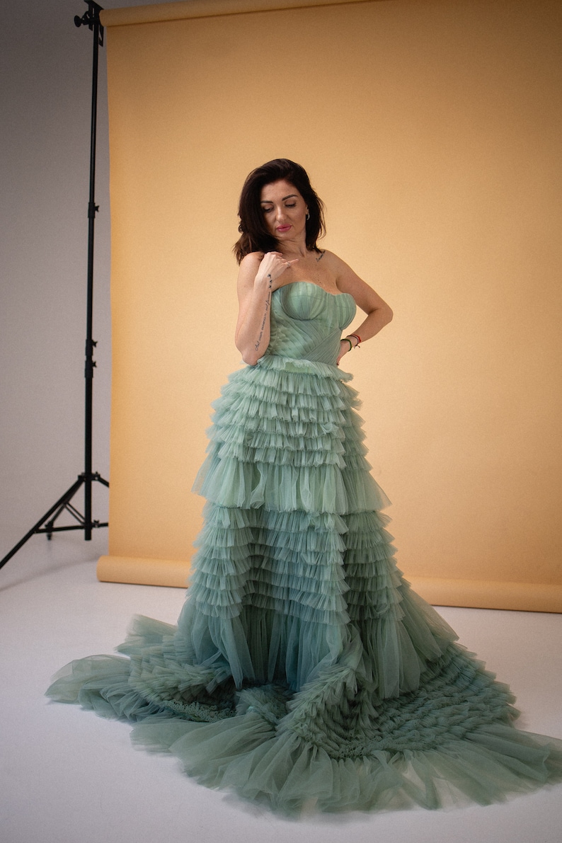 Pu&ograve; includere: Una donna indossa un abito di tulle verde salvia con pi&ugrave; strati di balze. L'abito ha un corpetto senza spalline e una gonna fluida.