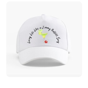 May include: White baseball cap featuring the text "Living life like a Jimmy Buffett Song" in black script. A margarita illustration with a lime and a red flower is also on the cap. The cap has a curved brim.