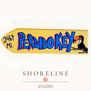 May include: A yellow wooden sign with the text "Perdido Key" in blue letters with a purple outline. The sign also features a cartoon toucan and the text "2467 Mi."