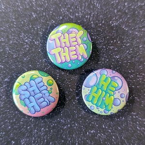 May include: Three round buttons with a bubble design. The buttons have the words "She Her", "He Him", and "They Them" written on them in a colourful, bubbly font.