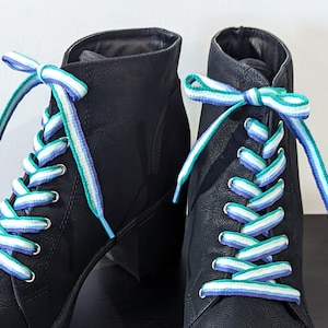 May include: Two black ankle boots with colourful striped laces. The laces display a pattern of white, teal, and purple stripes. The boots have silver eyelets. The image is set against a plain white background.