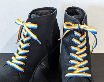 Puede incluir: Un par de botines negros con cordones amarillos, azules y blancos. Los cordones están atados en forma de cruz.