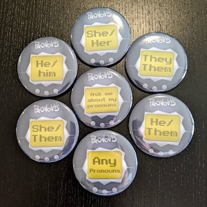 May include: Seven round, grey buttons with white text and yellow accents. Each button has the word "PRONOUNS" printed on it. The buttons display different pronoun options, including "She/Her", "He/Him", "They/Them", "She/Them", "He/Them", "Ask me about my pronouns", and "Any Pronouns".