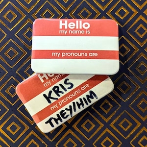 May include: Two red name tag badges with white text. The top badge says "Hello, my name is" and "my pronouns are". The bottom badge says "my name is KRIS" and "my pronouns are THEY/HIM".