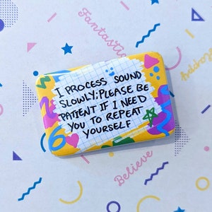 May include: A yellow button with a grid pattern and colourful accents. The text on the button reads: "I process sound slowly: Please be patient if I need you to repeat yourself."