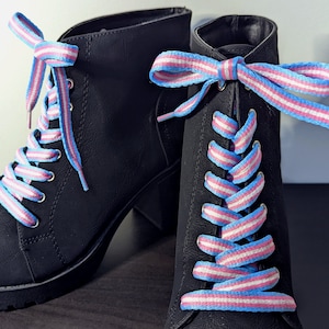May include: A pair of black ankle boots featuring pink, white, and light blue striped laces. The laces are tied in bows. The boots have a chunky heel and platform sole. The laces are threaded through the eyelets.